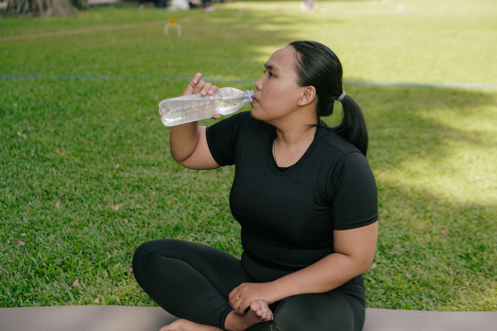 A woman in black workout clothes sits on a mat on the grass, drinking water after exercise